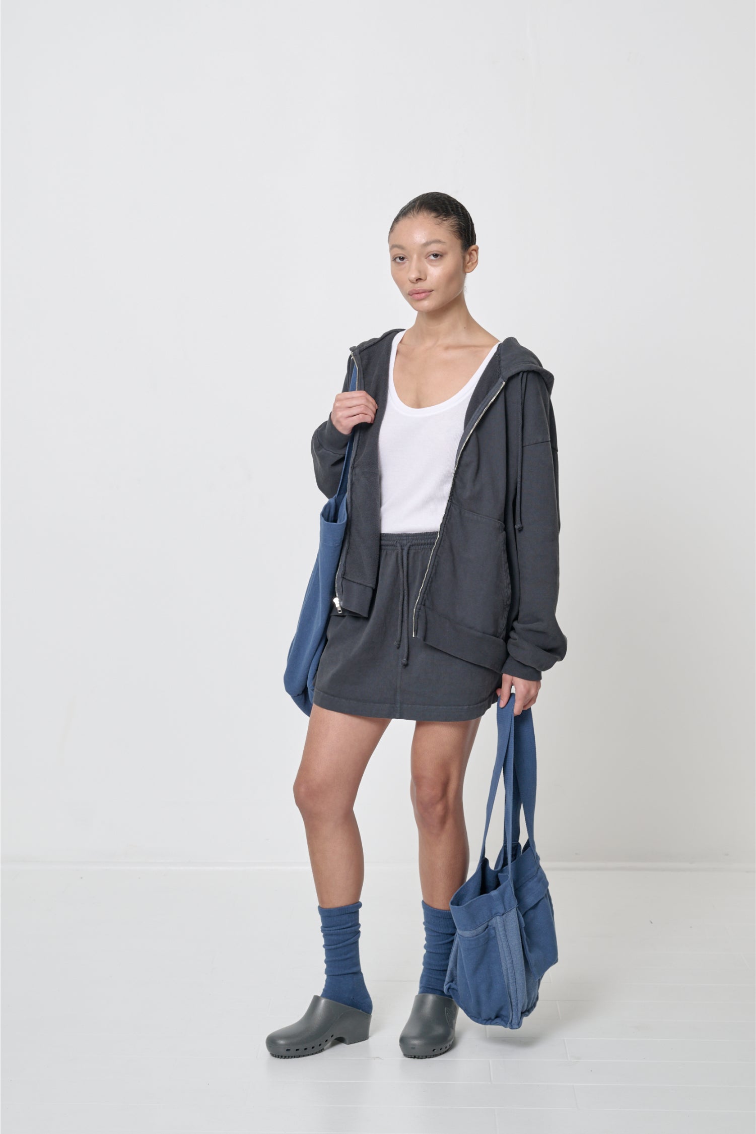 Front view: woman in an unzipped Slate Trash Zip Up Hoodie and Trash Skirt, carrying two blue tote bags.