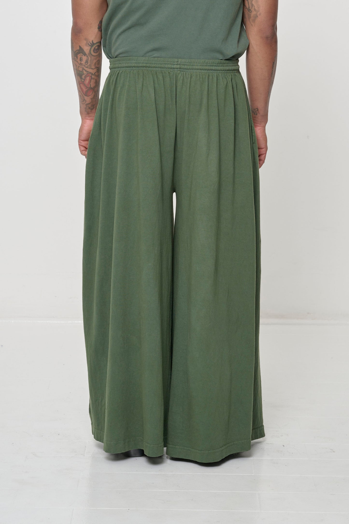 Back view: Lower 2/3 of a person wearing Pine Tubular Pants, flowing vertical fabric folds.