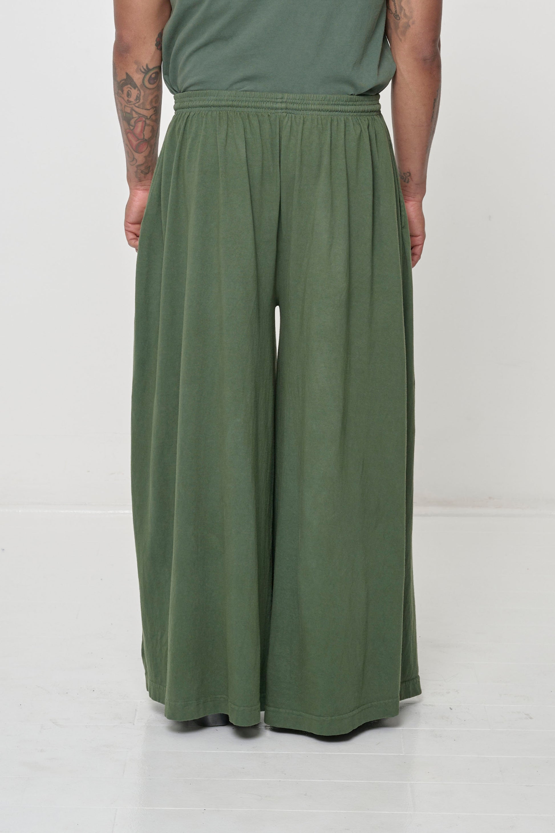 Back view: Lower 2/3 of a person wearing Pine Tubular Pants, flowing vertical fabric folds.