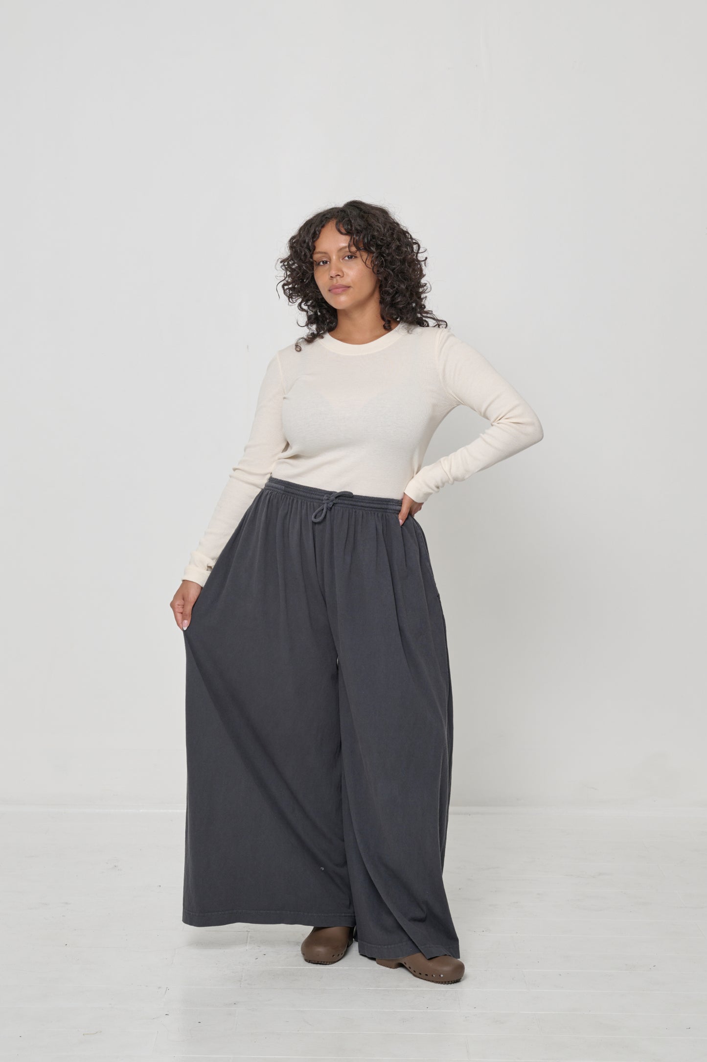 Woman in Tubular Pants in Slate, holding the side of one pant leg to display the wide legs.