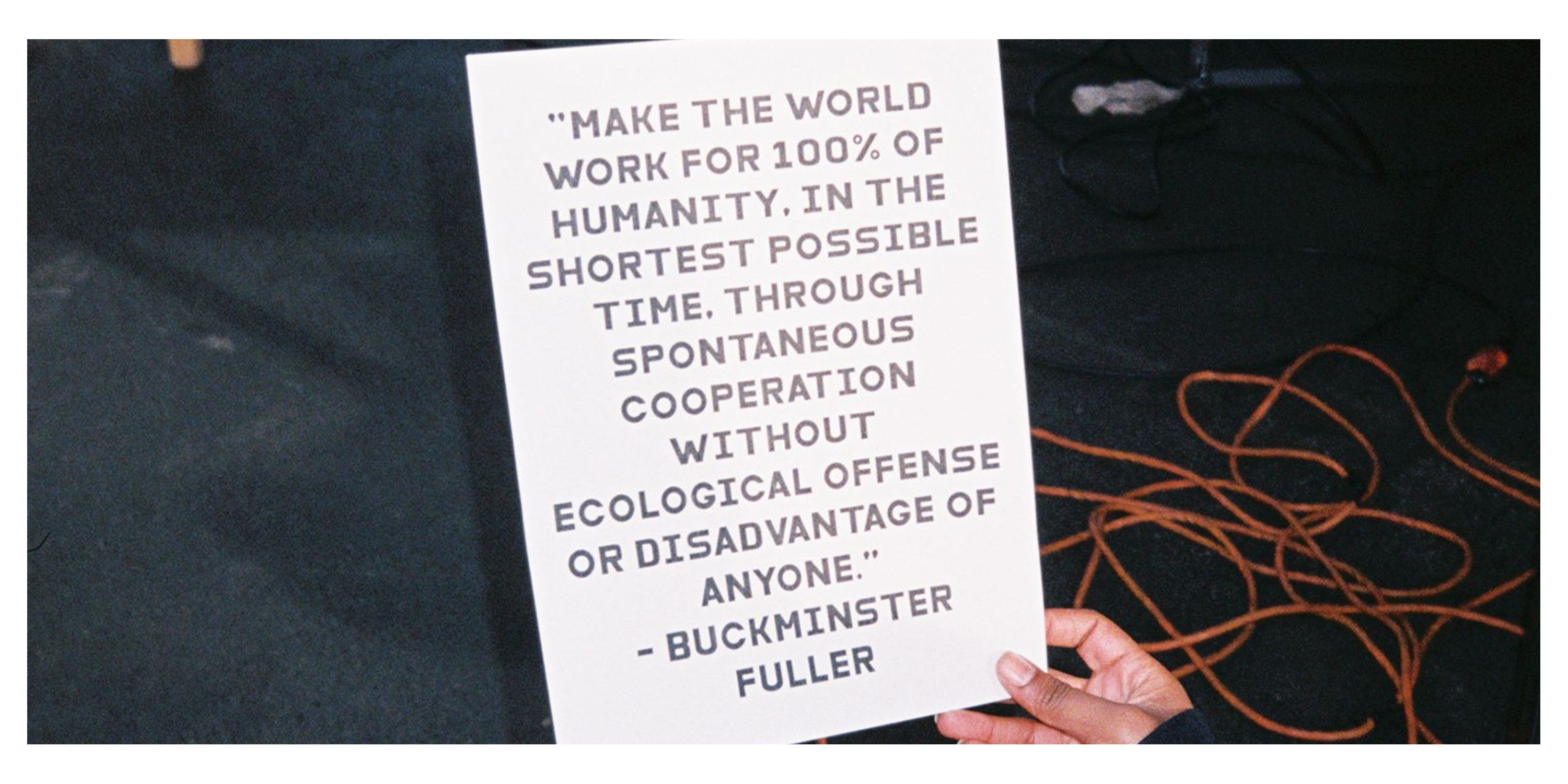Person holding a sign with a quote by Buckminster Fuller against a dark background