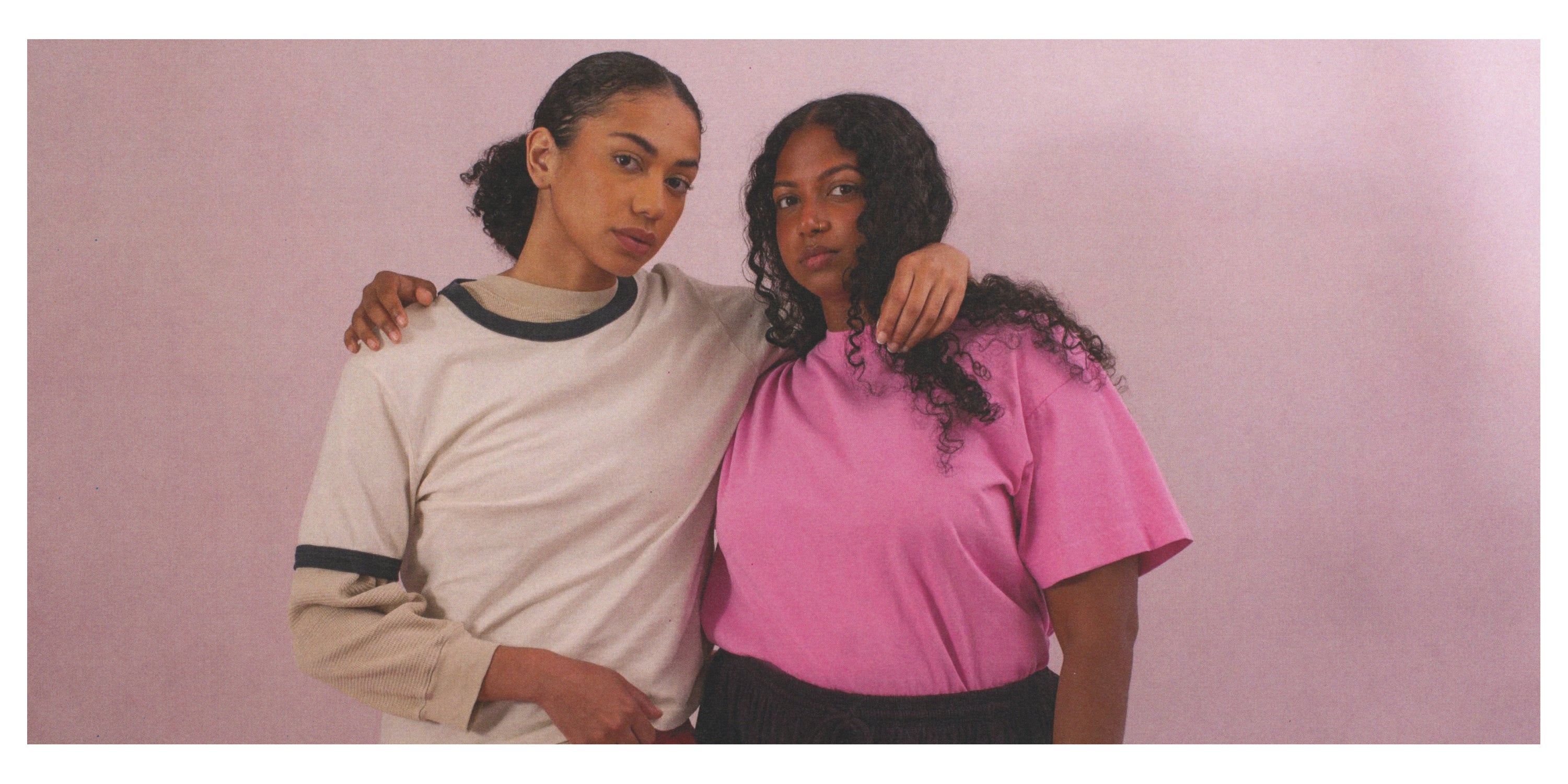 Two women standing close together against a pink background.