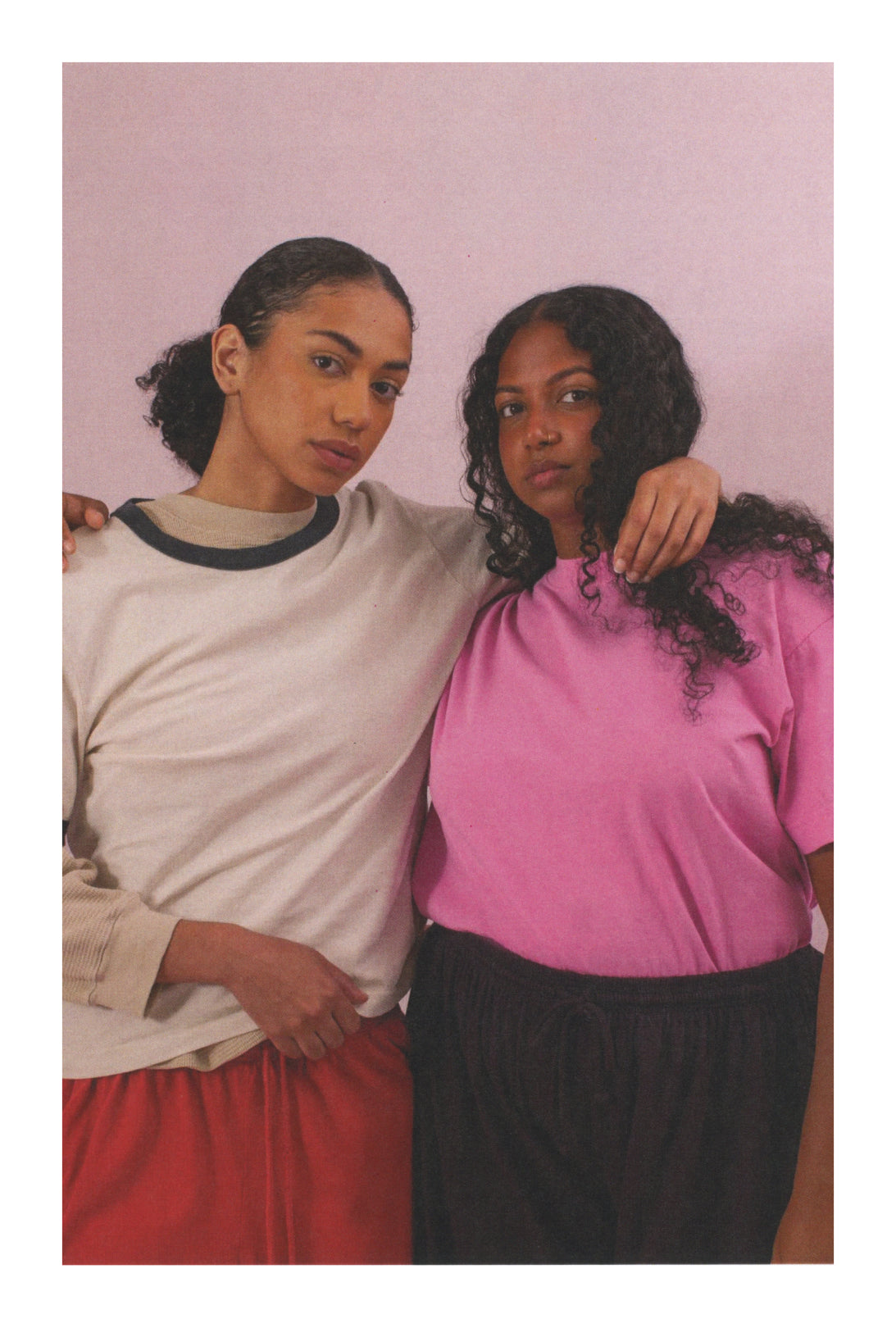 Two women standing close together against a pink background.