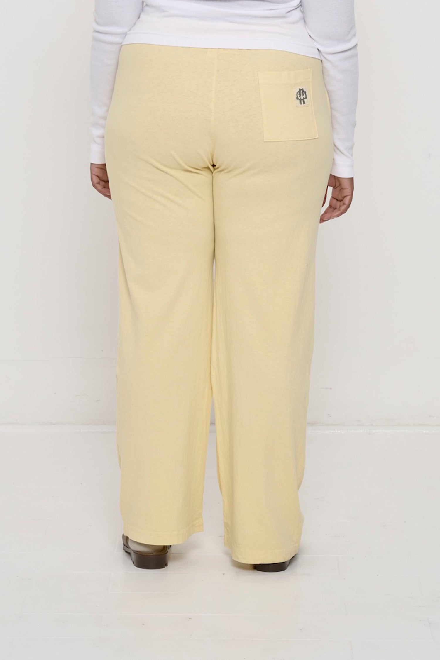 Back view, waist down: a person in French Butter Apartment Pants, the grey E.W logo printed on the right back pocket.