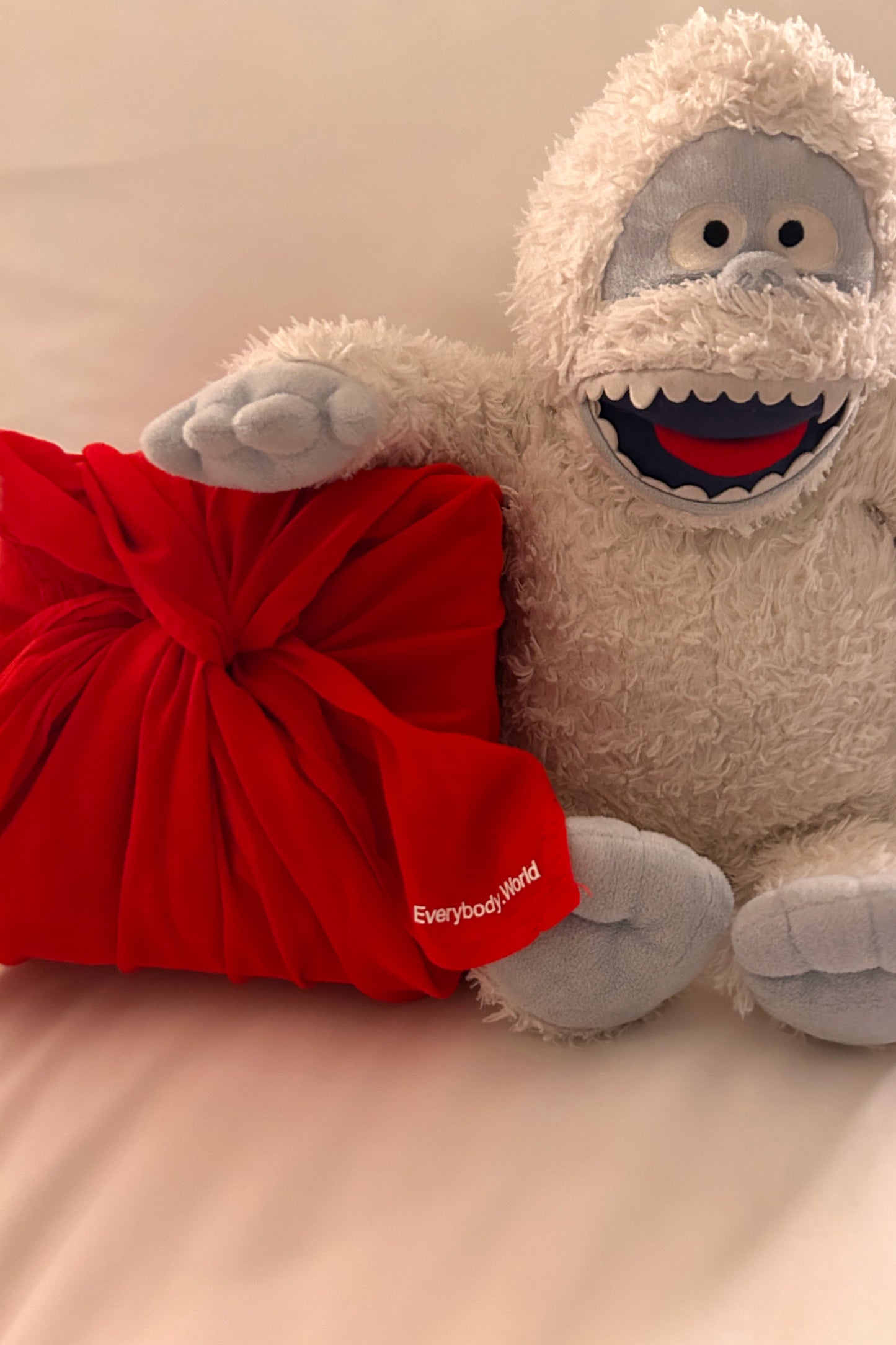 Plush abominable snowman toy with a red bag labeled 'Everybody'World' on a beige background