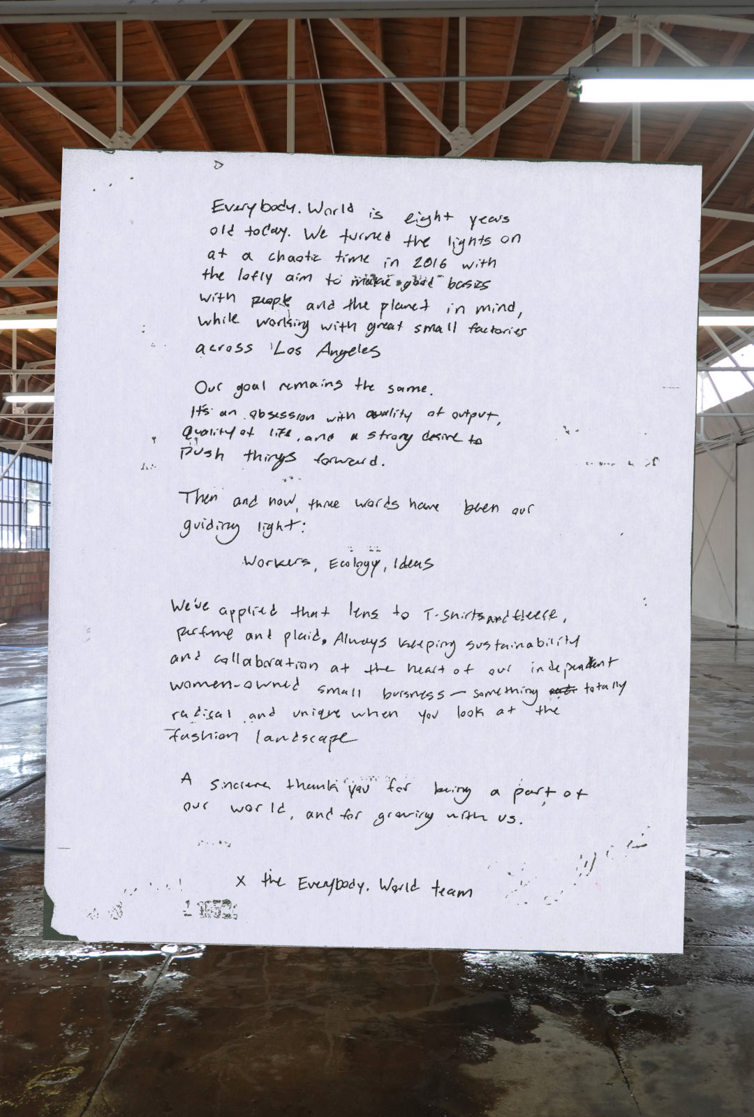 Handwritten note on white paper in a warehouse with high ceilings and wooden beams. The note celebrates Everybody.World's eighth anniversary, reflecting on its 2016 founding with a mission to create sustainable, quality basics with small LA factories. It emphasizes 'Workers, Ecology, Ideas' and highlights the brand's commitment to sustainability as a woman-owned business. It ends with a thank you, signed 'x the Everybody.World team.