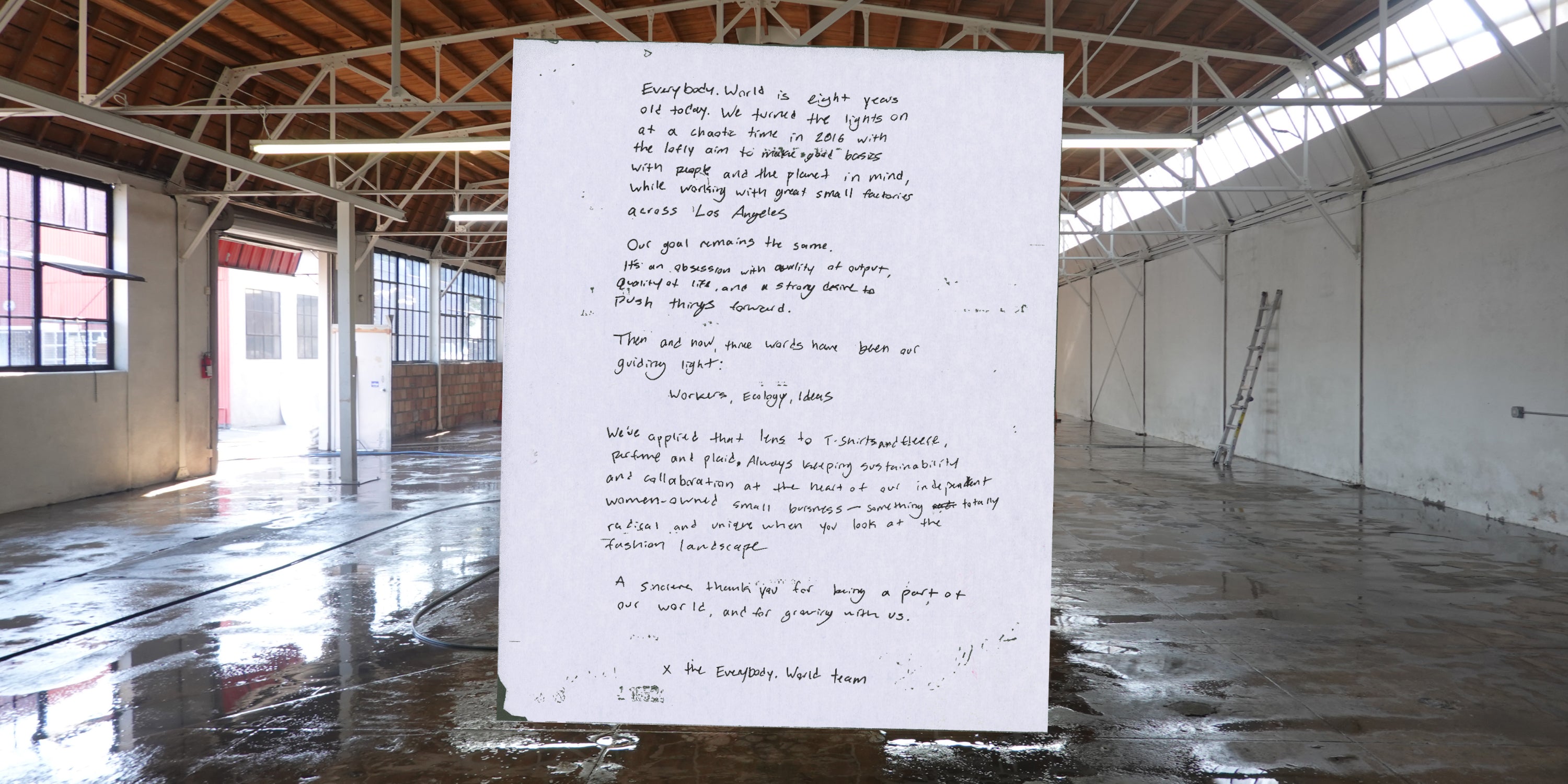 Handwritten note on white paper in a warehouse with high ceilings and wooden beams. The note celebrates Everybody.World's eighth anniversary, reflecting on its 2016 founding with a mission to create sustainable, quality basics with small LA factories. It emphasizes 'Workers, Ecology, Ideas' and highlights the brand's commitment to sustainability as a woman-owned business. It ends with a thank you, signed 'x the Everybody.World team.