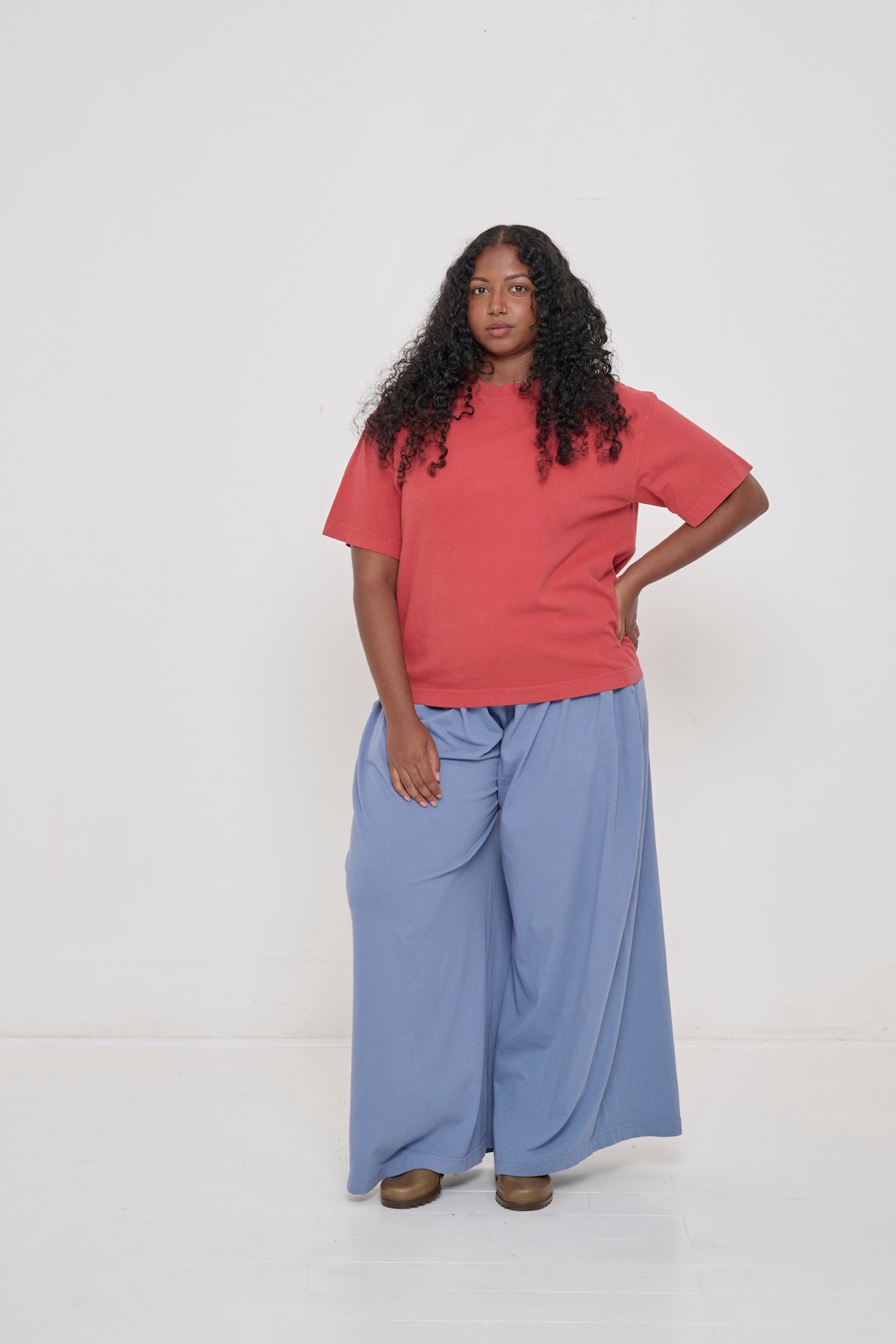 A woman wearing a Cedar (orange-red) Boxier Trash Tee and Periwinkle Tubular Pants, with a hand on her hip.