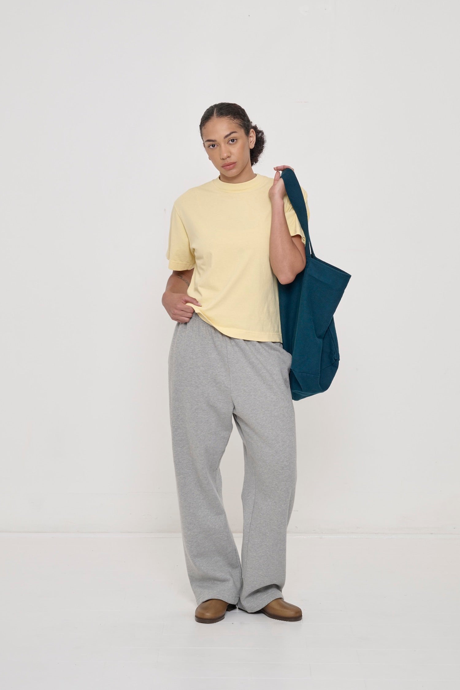 Woman in a French Butter Boxier Trash Tee and Heather Sweatpants, holding a Juniper (dark teal) Big Tote over her shoulder.