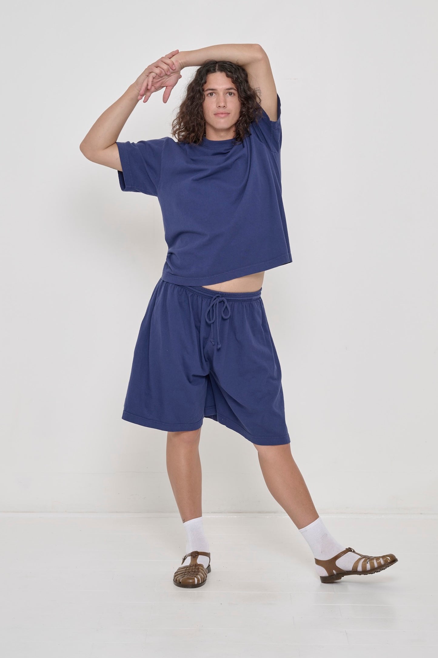 A person in matching Washed Navy Boxier Trash Tee, and Long Tubular Shorts. Arms atop head, raising tee hem just above the waist.