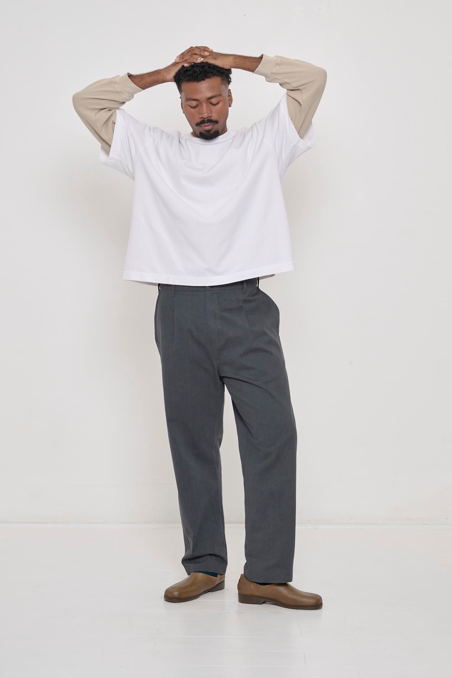 A man in a White Boxier Trash Tee, and Cotton Twill Life Uniform Pants. Hands atop head, which raises tee hem to his waist.