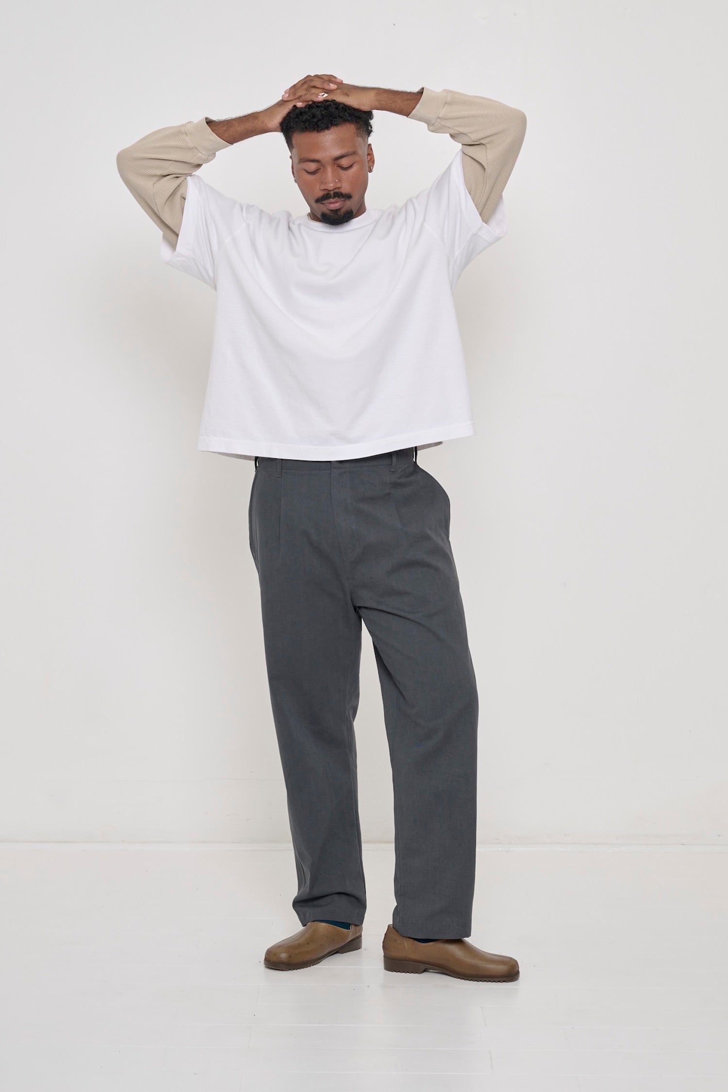 A man in a White Boxier Trash Tee, and Cotton Twill Life Uniform Pants. Hands atop head, which raises tee hem to his waist.