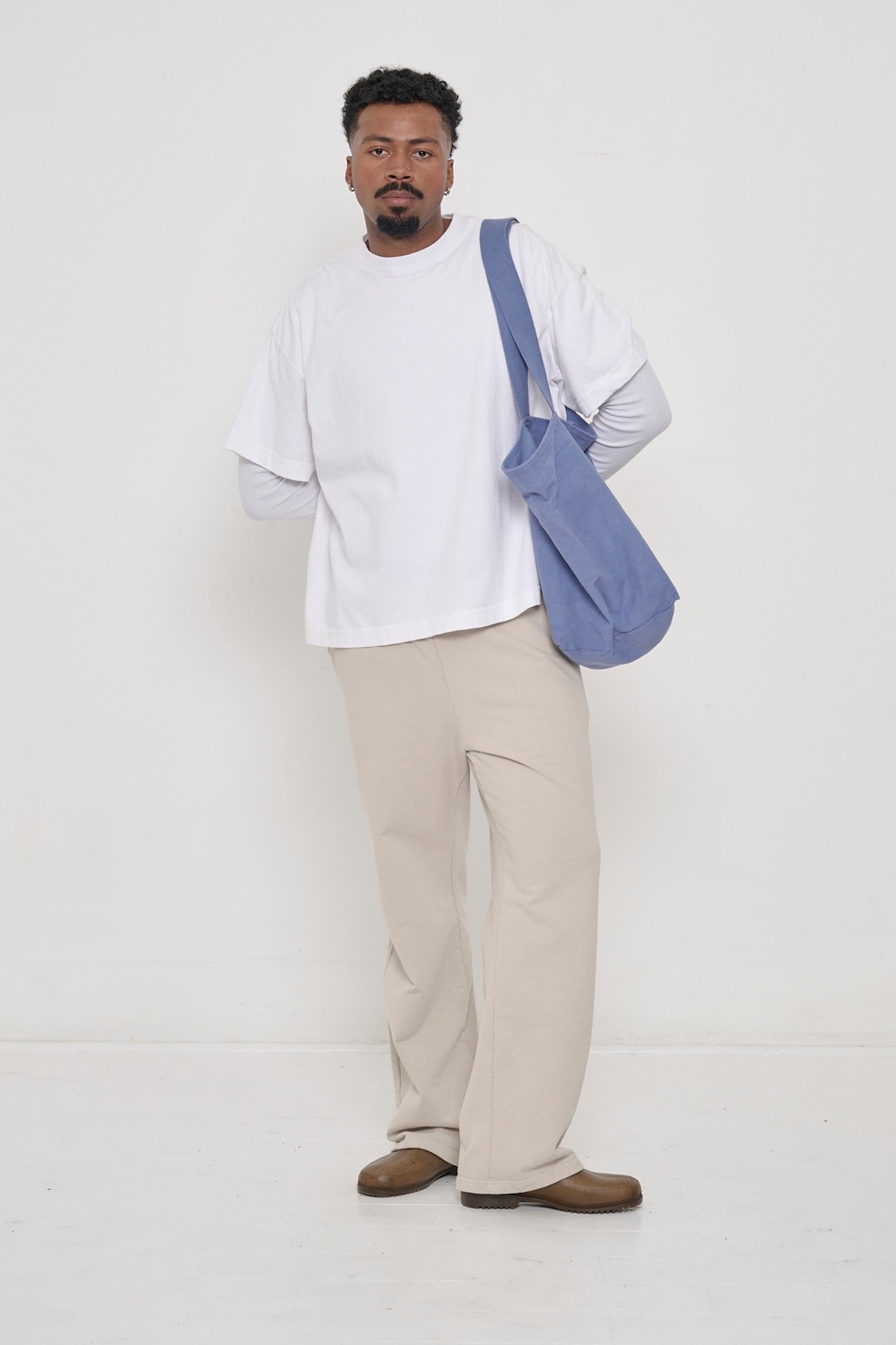 A man in a White Heavyweight Boxier Trash Tee with Oat Trash Fleece Apartment Pants and a Big Tote over his left shoulder.