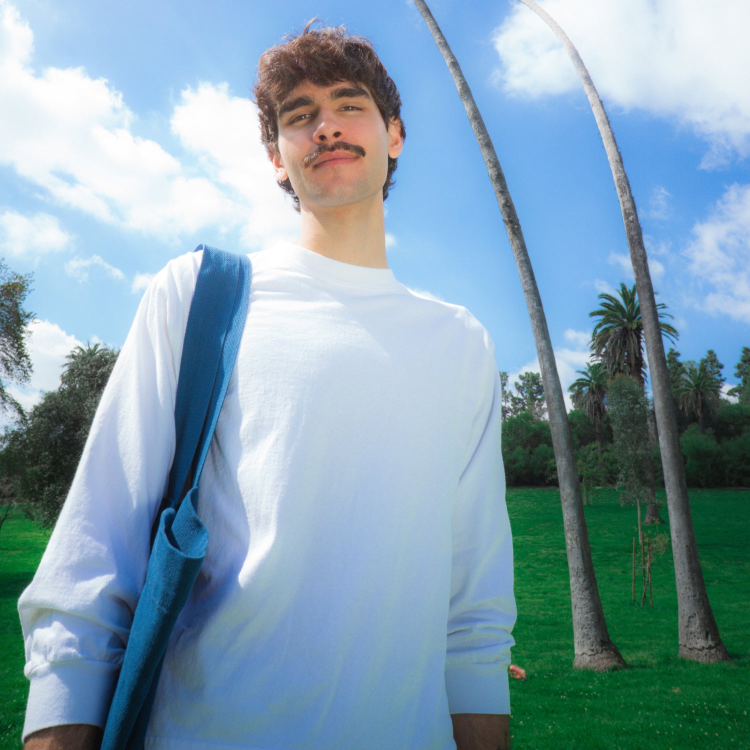 A model wears a White Long Sleeve Boxier Trash Tee and holds a Tote over his shoulder, two palm trees are in the background