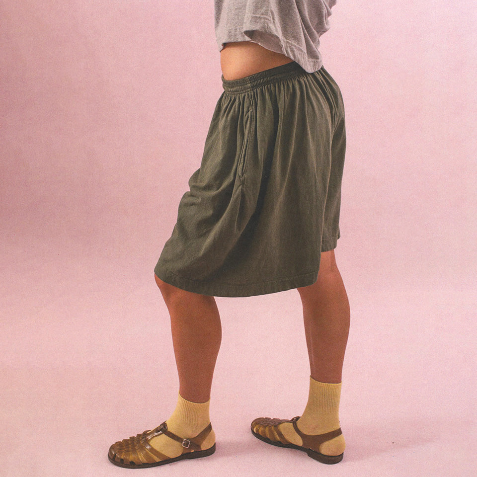 Bottom half of a person wearing Long Tubular Shorts in Pine, a Heather Boxier Tee that is showing some midriff and Spring Socks in Butter Yellow.