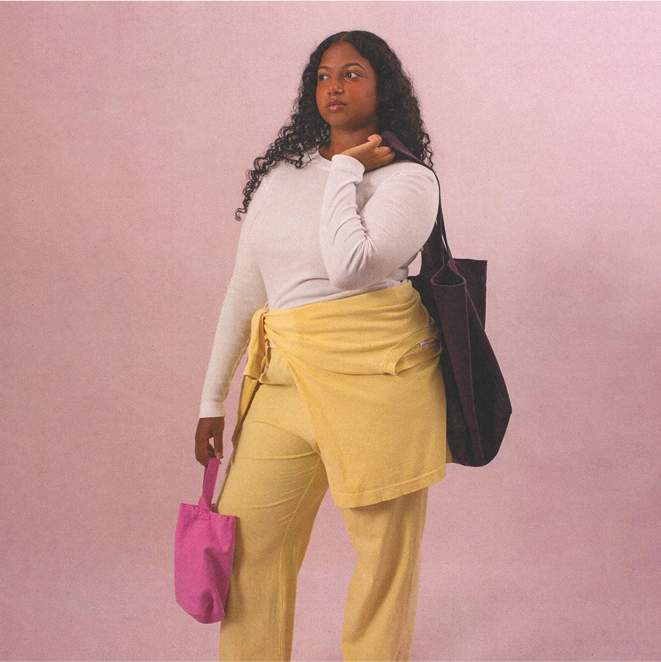 Woman wearing a Rib Long Sleeve and Apartment Pants, holding a Pixie Loop Tote and a Mulberry Big Tote, against a pink background.