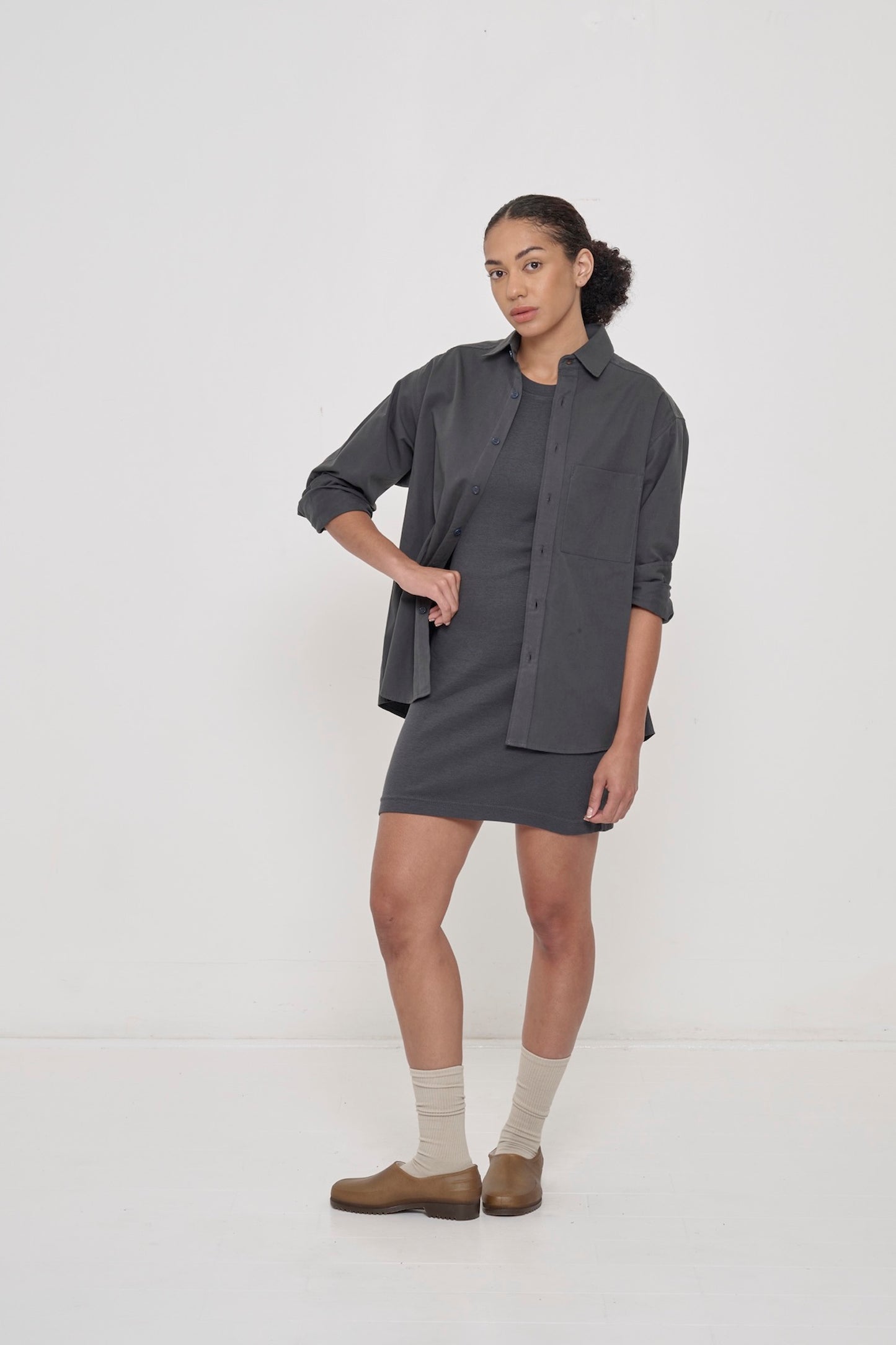 A woman with one hand on her hip, wearing a Slate Rib Dress under an open Slate Peach Fuzz Twill Button-Up Shirt.