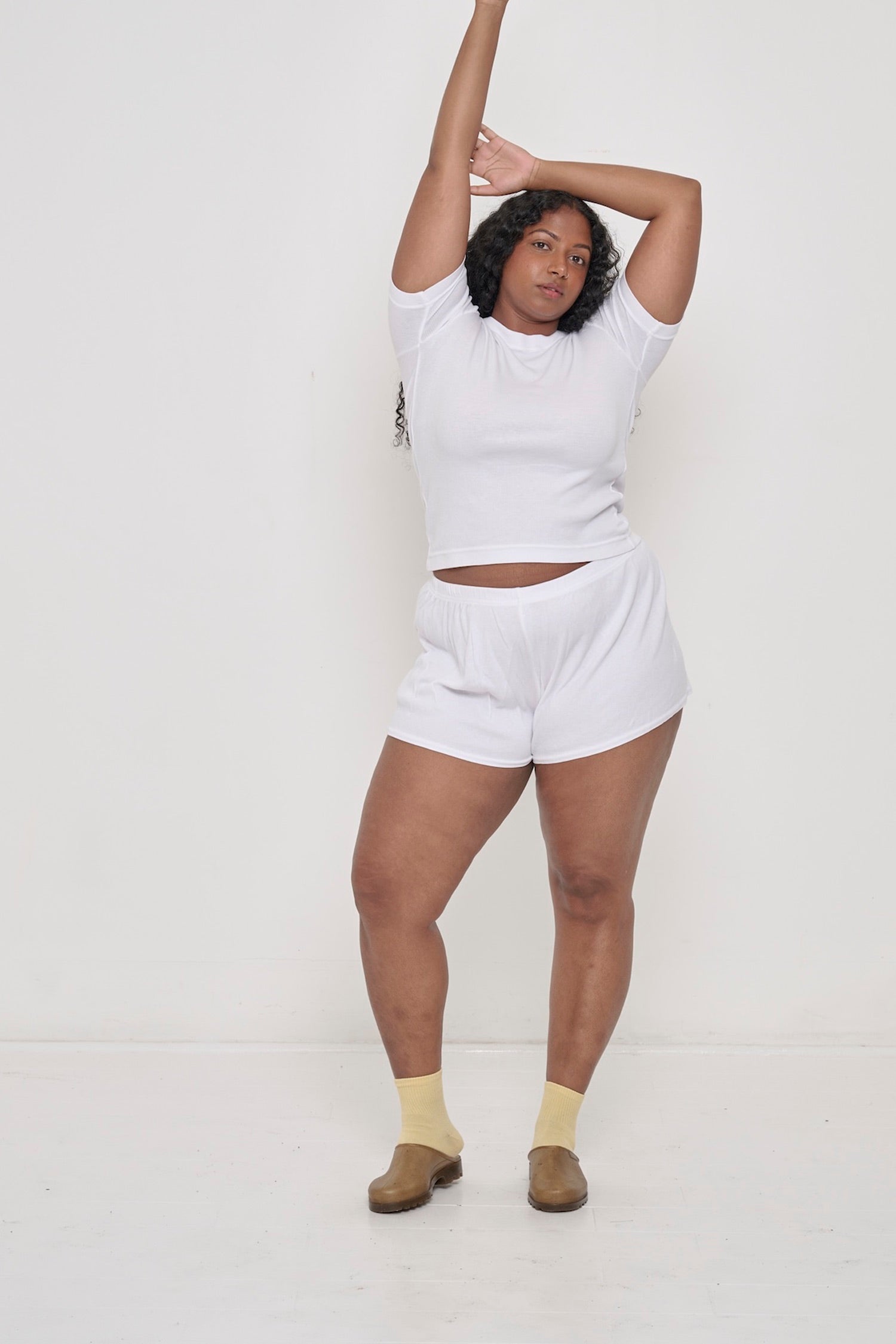 A woman in matching White Rib Short Sleeve and Rib Shorts. Arms raise up, raising shirt to expose an inch of her torso.