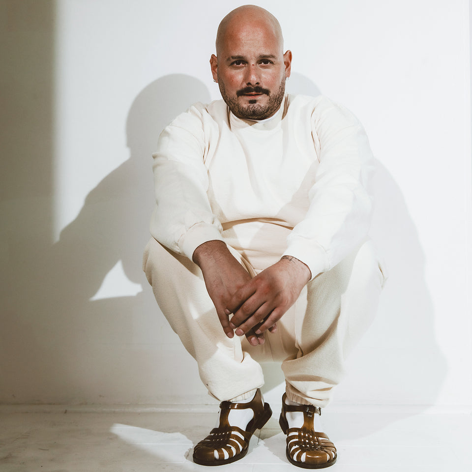 Man in Trash Crewneck and Trash Sweatpants both in Scour, crouched in a white room.