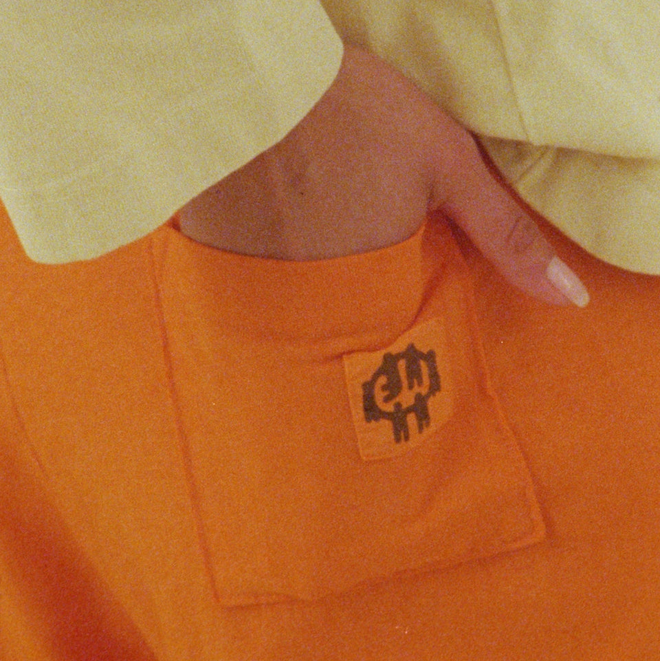 Close-up of the back pocket with Everybody World logo of the Apartment Pants in Persimmon, with the model's hand partially in the pocket.