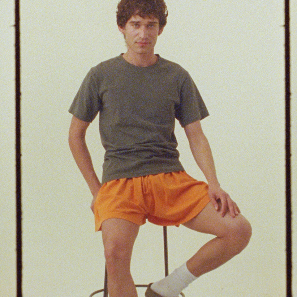 Person wearing a Short Sleeve Trash Tee in Pine and Tubular Shorts in Persimmon, they are sitting on a stool against a plain background