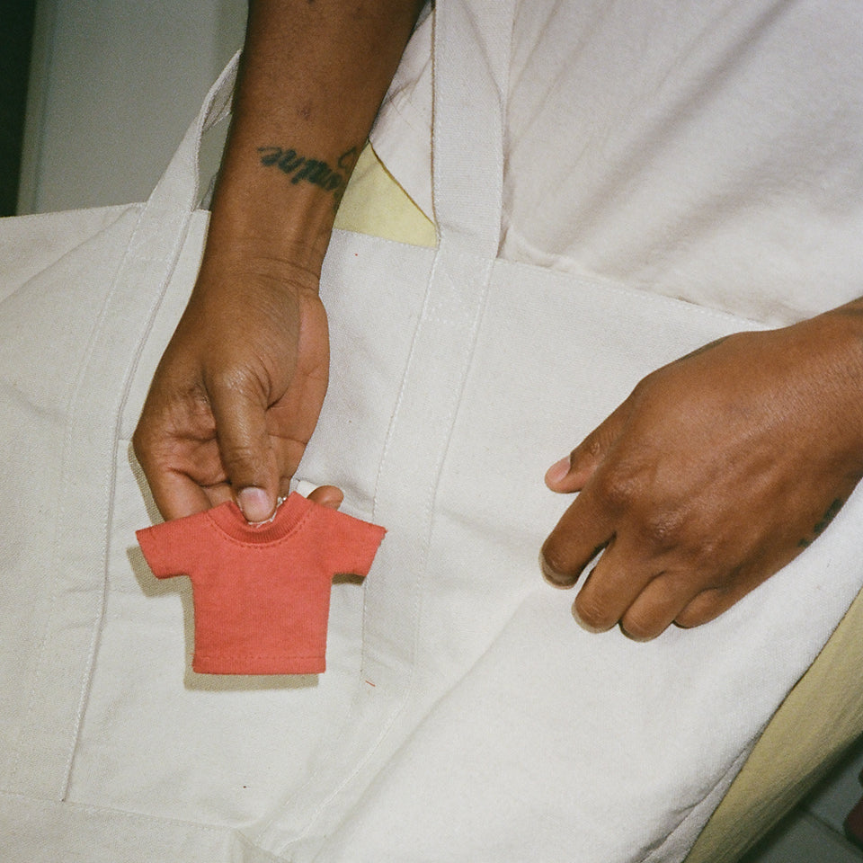 Person holding a mini Trash Tee charm keychain that is attached to the Overnight Tote, in color Natural. 