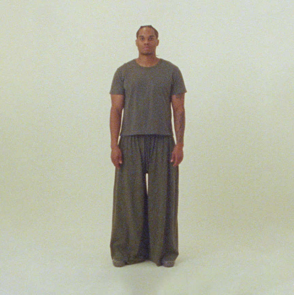Person wearing a Tailored Trash Tee and Tubular Pants in the color Pine, standing against and plain background. 