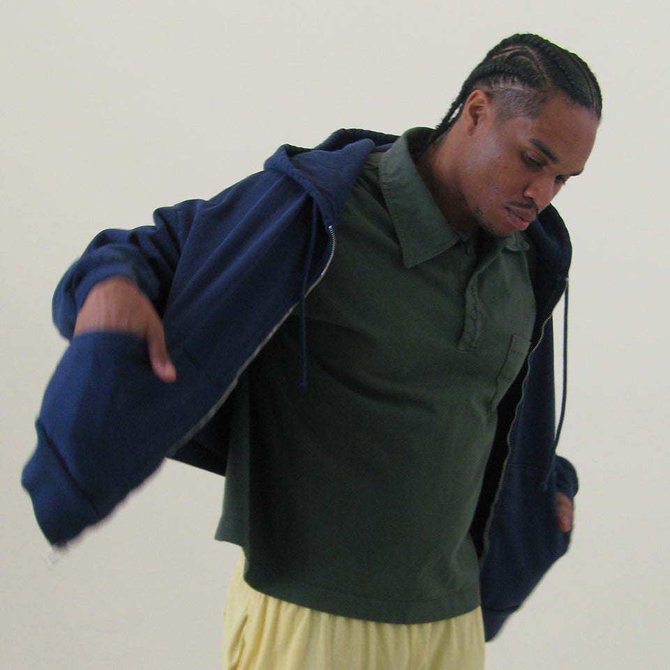Man pulling on a Washed Navy Trash Zip Up Hoodie over a Pine Trash Polo, standing against a plain background
