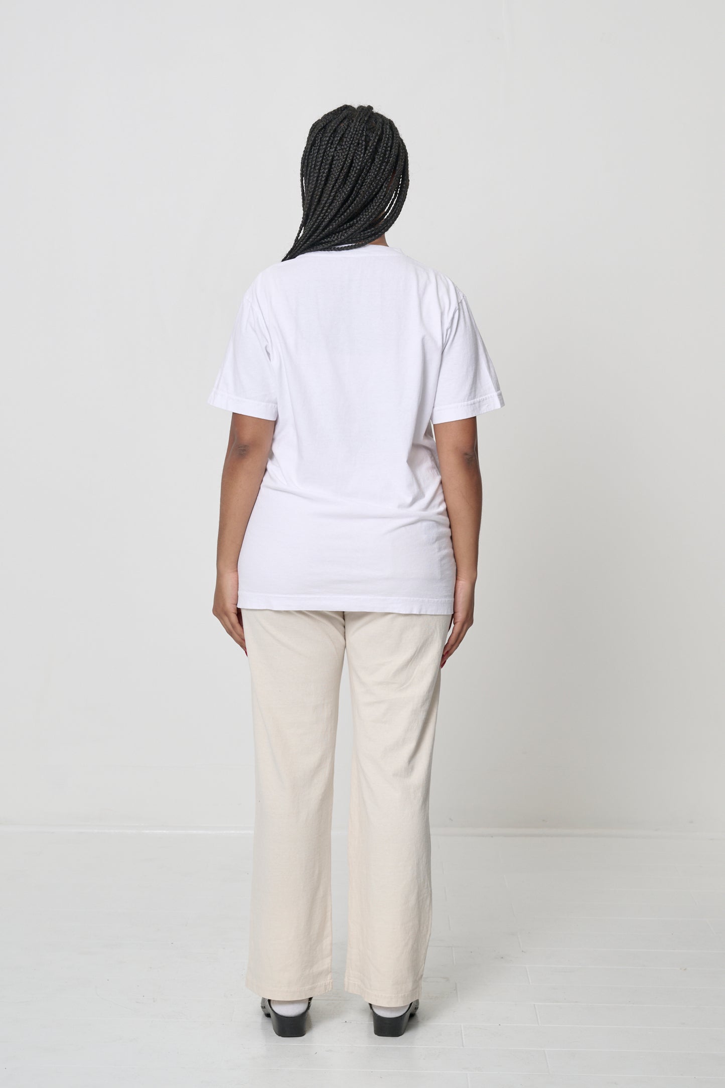 Back view: a woman in a White Short Sleeve Trash Tee, hair is pulled to the side showing the back neckline. 