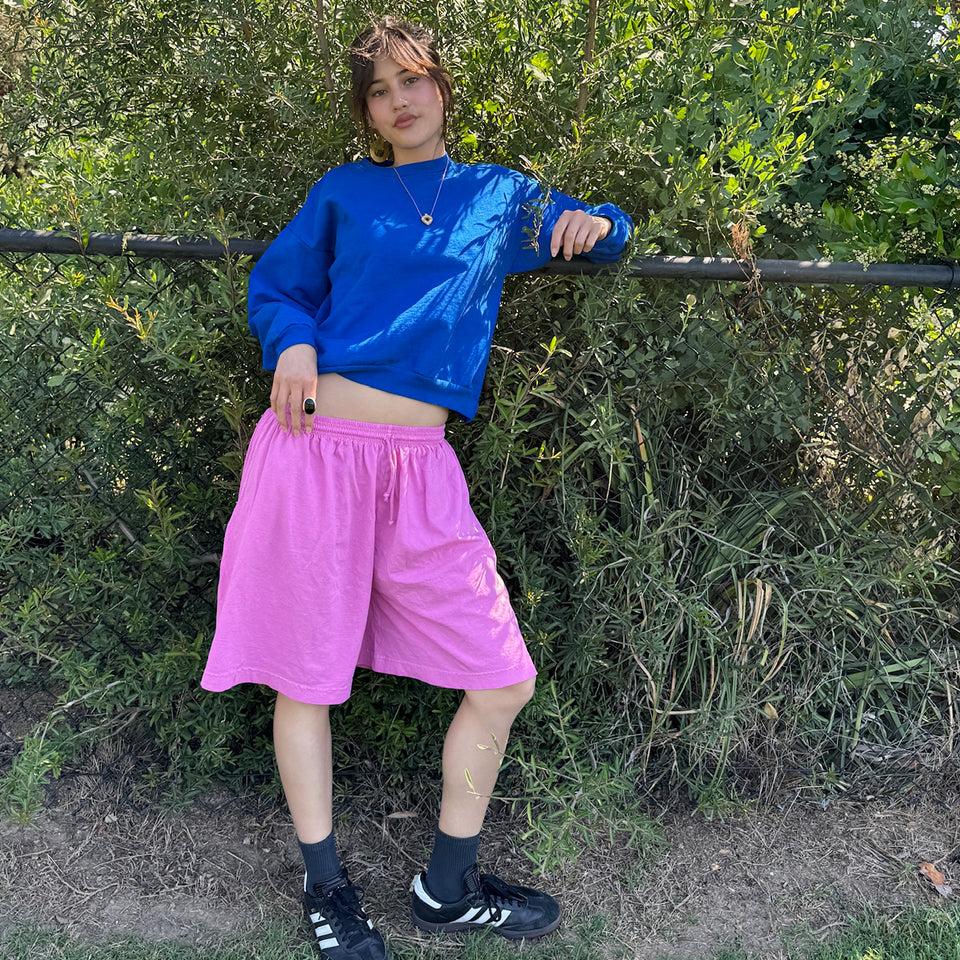 Woman leans against fence outside, wearing Long Tubular Shorts in pixie and Trash Crewneck in Lapis.