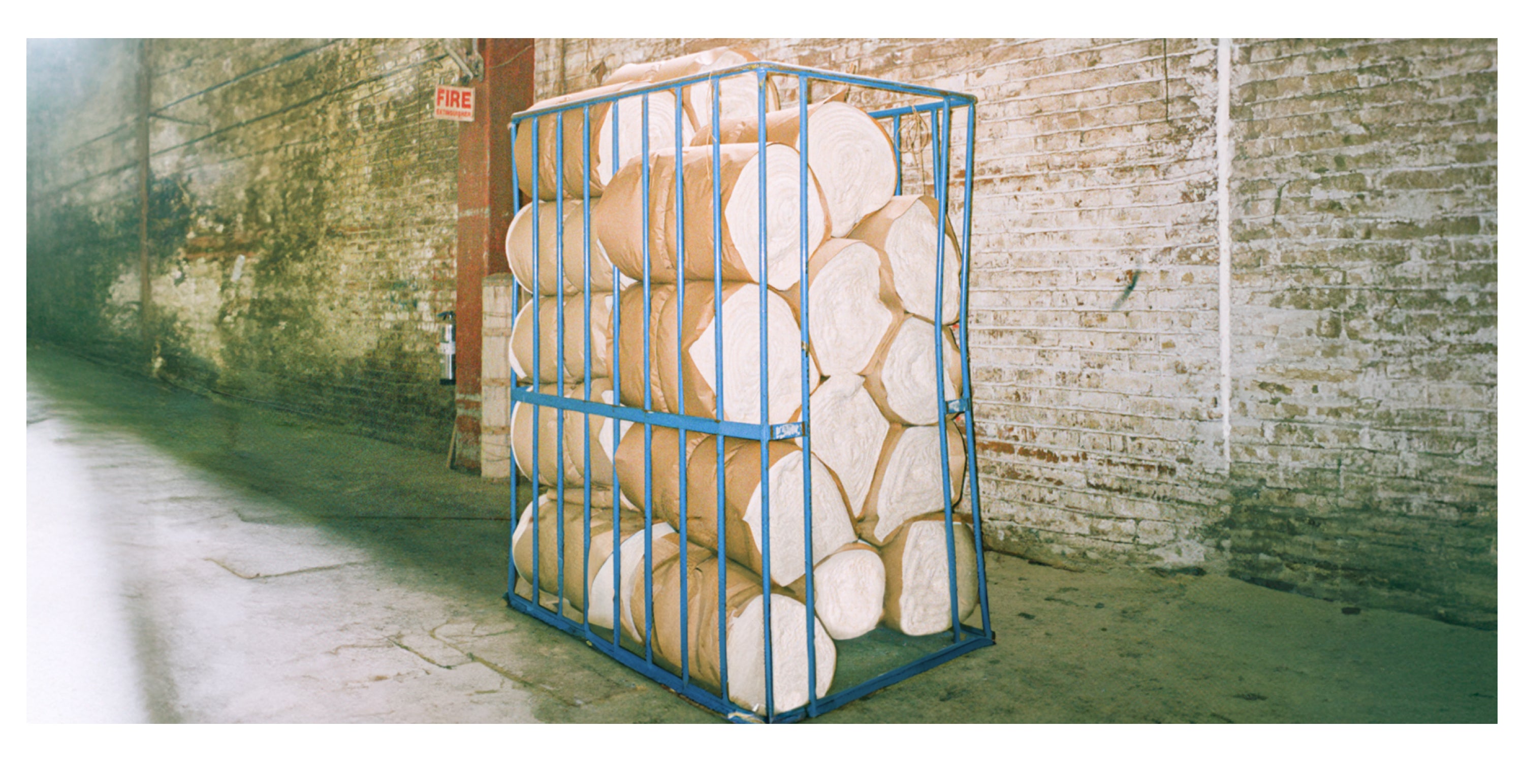 A blue metal frame containing compact rolls of brown paper-wrapped material, placed in an industrial warehouse with exposed brick walls.