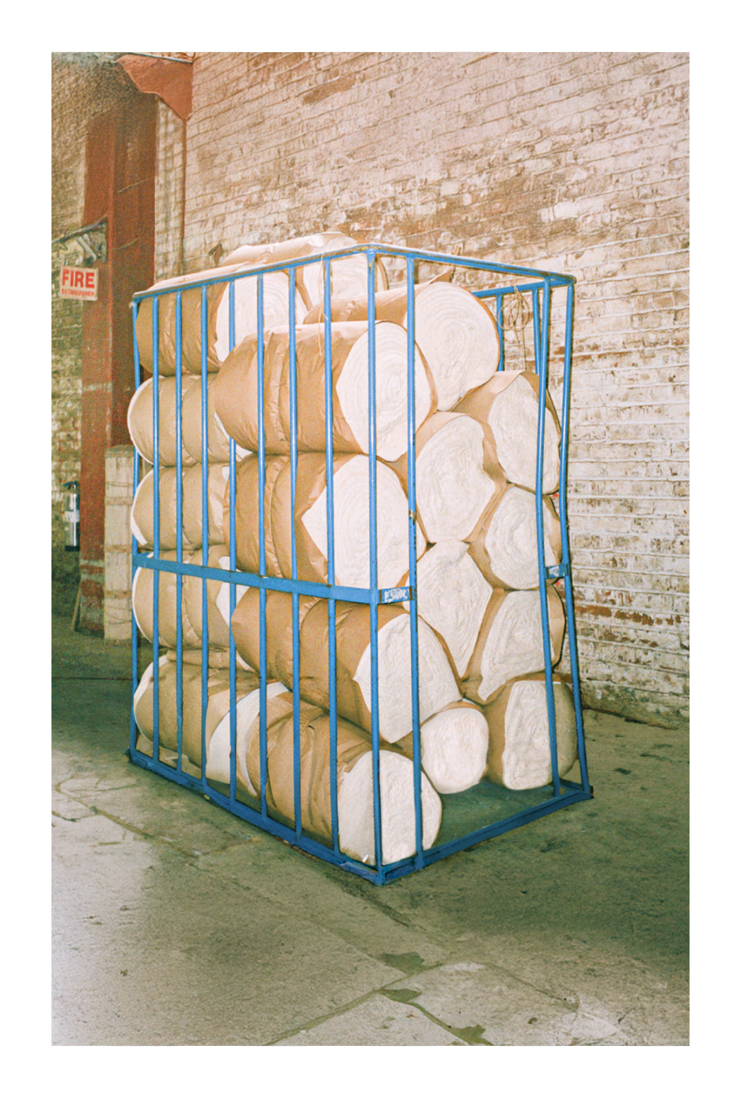 A blue metal frame containing compact rolls of brown paper-wrapped material, placed in an industrial warehouse with exposed brick walls.