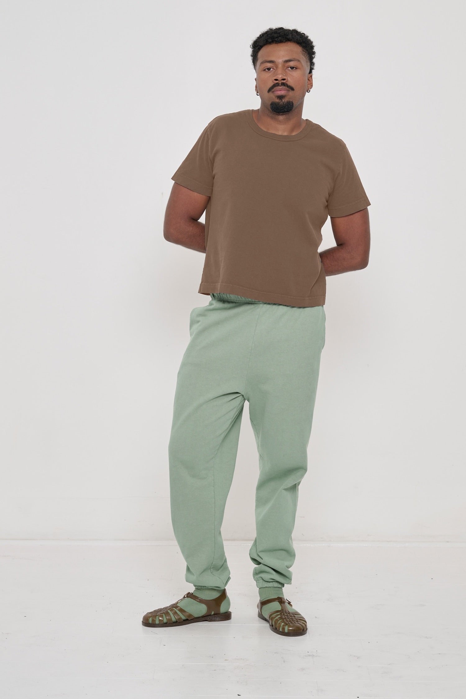Man in Dry Soil Tailored Trash Tee and Wasabi Trash Sweatpants, both hands behind back, showing shirt's slim sleeves.