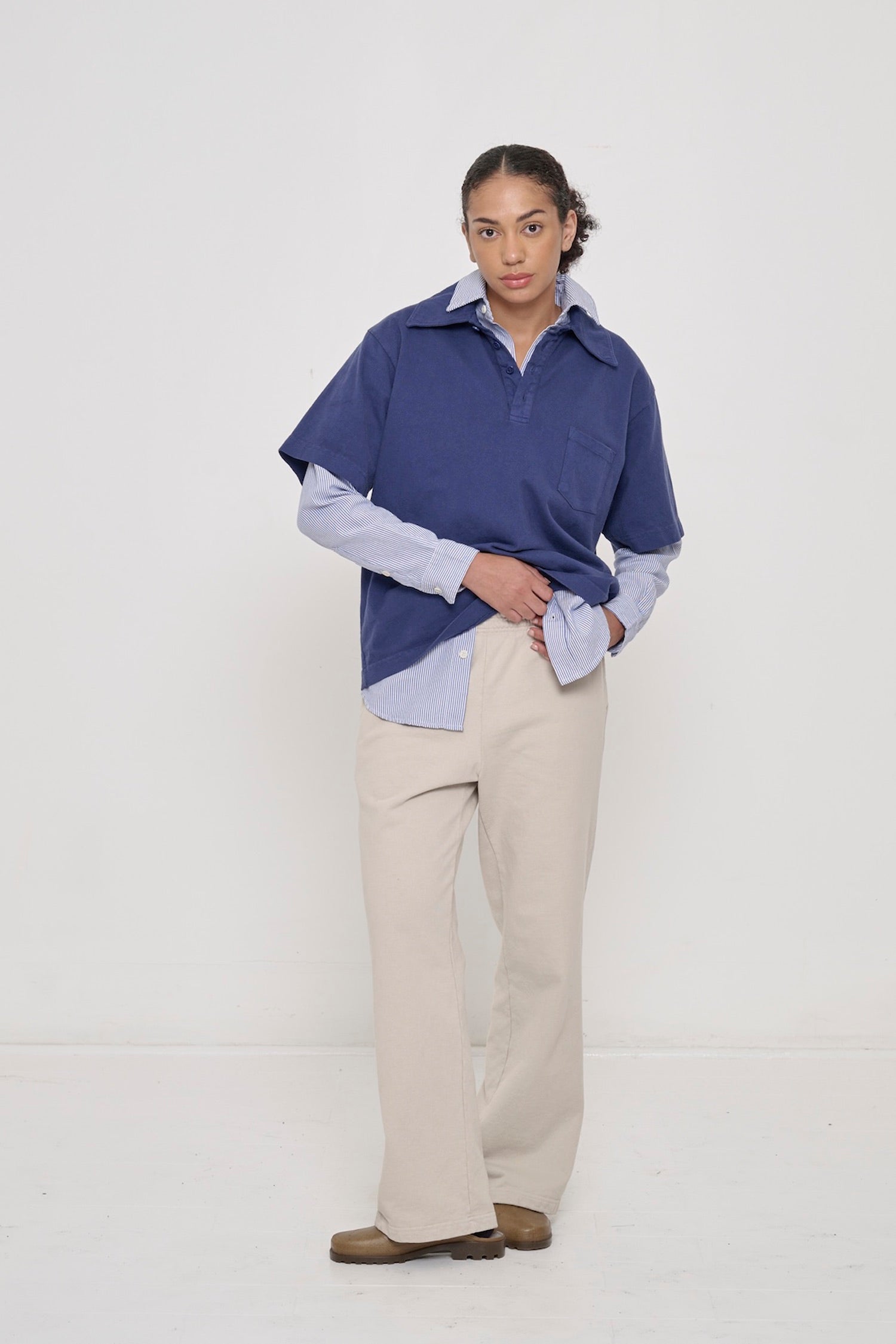 A woman wearing Trash Fleece Apartment Pants in Oat with layered Trash Polo in Washed Navy and striped button up shirt under.
