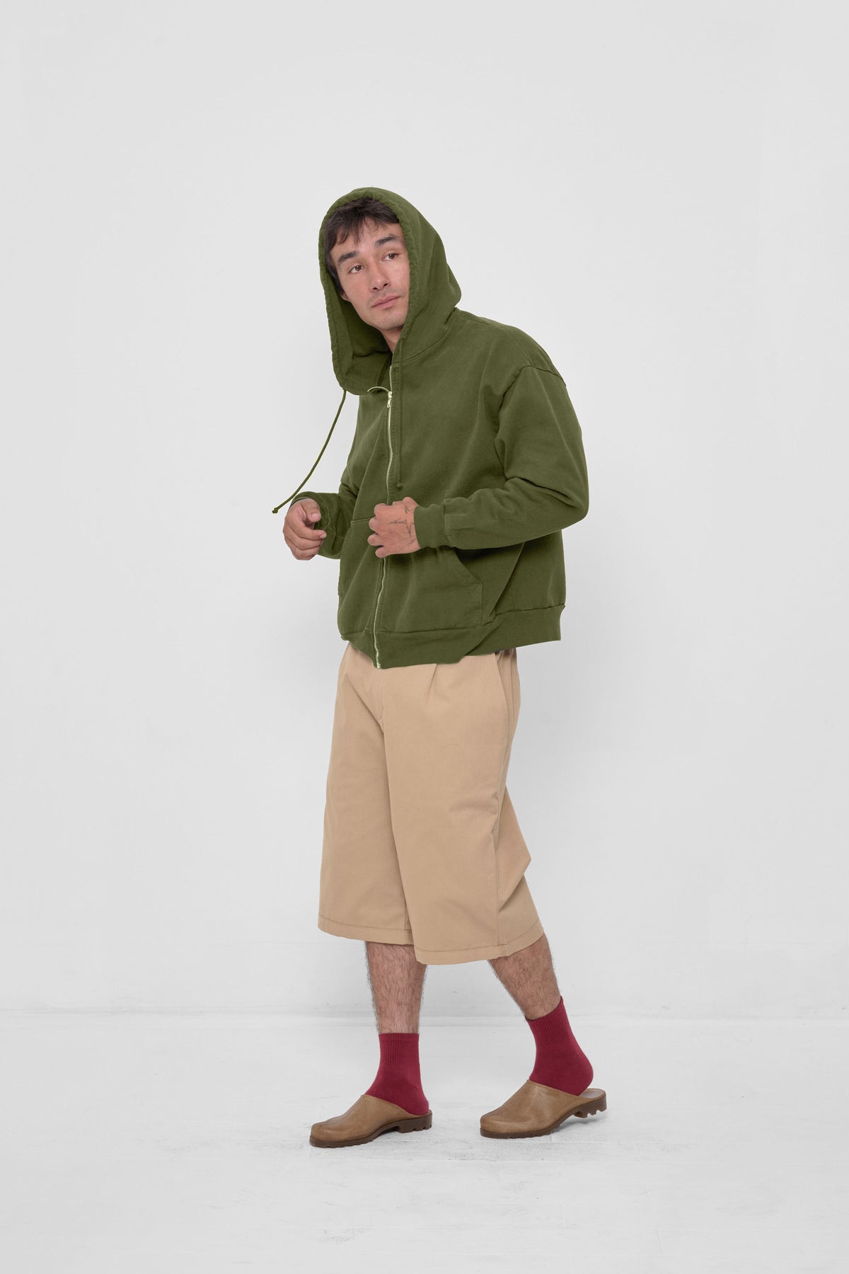 Trash Zip Up Hoodie in Olive – front fulll body view on model, styled with Sand Storm Bermuda Shorts, Garnet Squishy Socks, and Plasticana Mules.