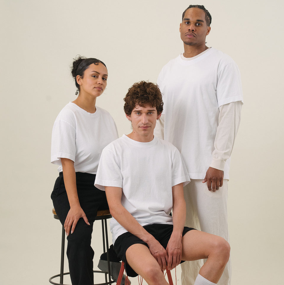 Three models against a white background. The model on the left is sitting and wearing a white Short Sleeve Trash Tee and black pants. The model in the middle is sitting and wearing a white Short Sleeve Trash Tee and black shorts. The model on the right is standing and wearing a white Short Sleeve Trash Tee over a Long Sleeve Trash Tee, with white pants.