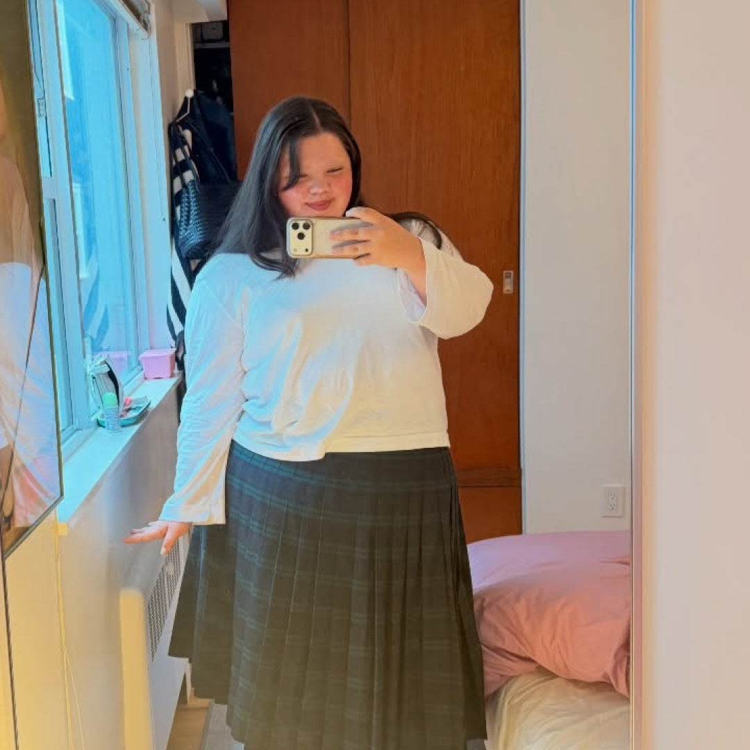 A person taking a mirror selfie in a bedroom, wearing a White Straight Sleeve Tailored Trash Tee and a plaid skirt. 