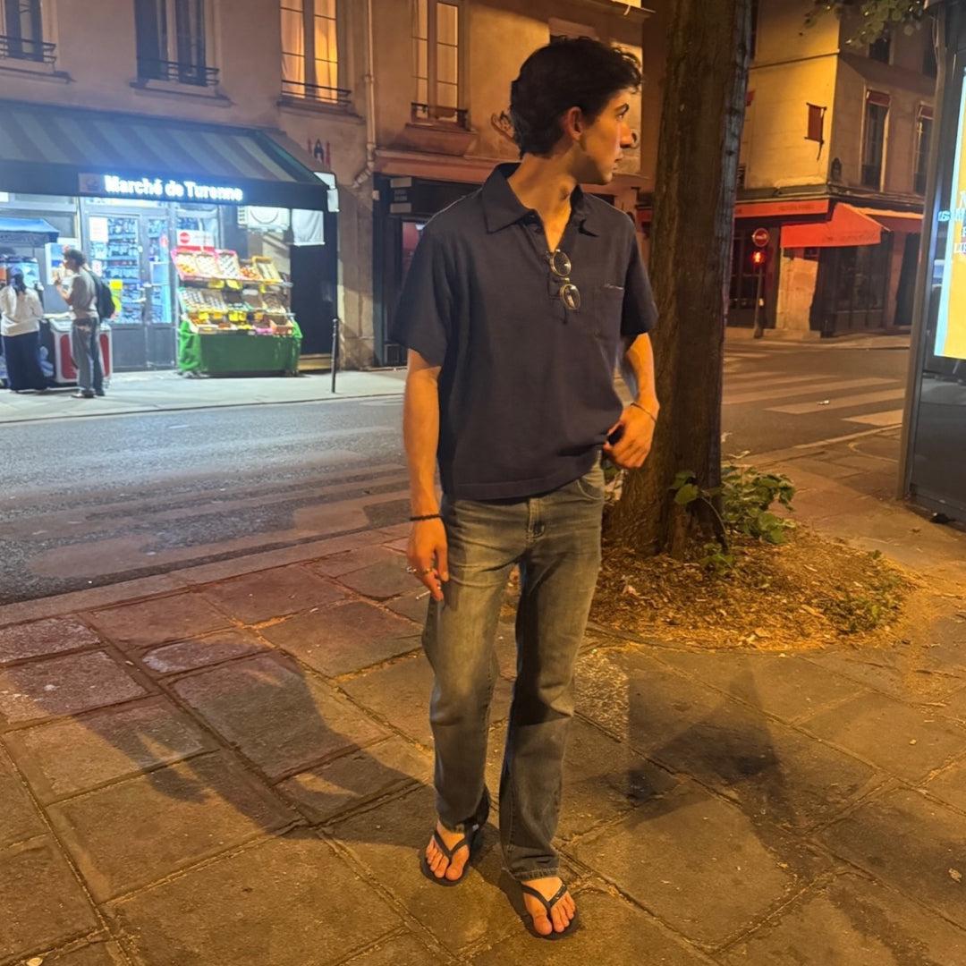A man wearing a Washed Navy Trash Polo, gray jeans, and black flip-flops standing on a city sidewalk at night.