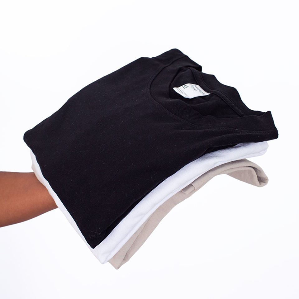 Hand holding three folded Trash Tees in Black, White, and Oat. 